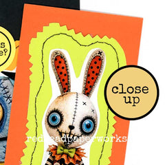 A close-up of a handmade mixed media Halloween Card featuring a stitched scary bunny face with blue eyes, red polka-dot ears, and a green bow tie, set against layered orange and yellow paper shapes.